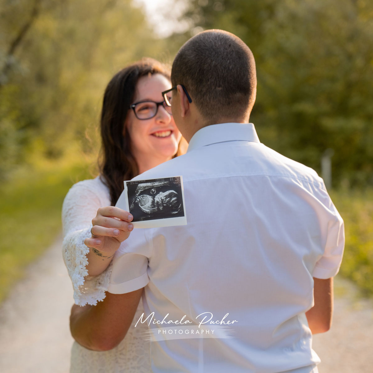Schwangere Frau umarmt ihren Partner von hinten und hält ein Ultraschallbild in die Kamera; beide lachen im warmen Abendlicht auf einem Waldweg.